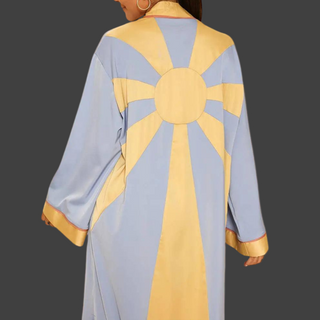 Back view of a woman wearing a flowing, silky blue and yellow kimono with a bold rising sun design, featuring long sleeves, gold trim, and side slits — styled for resort or statement wear.
