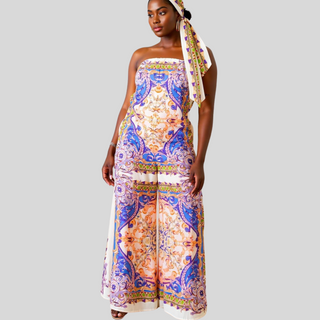 Model wearing the Sunny Day in Capri Jumpsuit, a strapless printed wide-leg jumpsuit with a removable tie belt, styled in warm orange, lilac, and green tones. Lightweight and perfect for summer vacations.