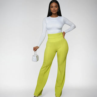 A stylish woman poses confidently in bright lime green high-waisted pants with a wide-leg fit, paired with a fitted long-sleeve white top and a small quilted white handbag. The pants feature vertical seam detailing, creating a sleek and elongating silhouette.